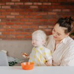 a mother smiles at her albino toddler indoors against a brick wall.