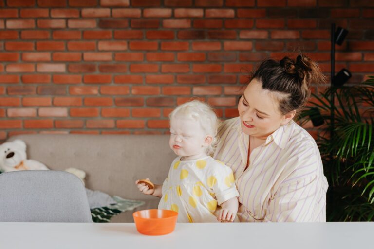 a mother smiles at her albino toddler indoors against a brick wall.