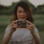 a smiling pregnant woman holds an ultrasound photo outdoors, symbolizing joy and anticipation.