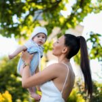 a joyful mother holding her baby under a green leafy tree on a sunny day.